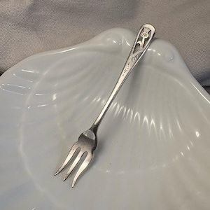 Op de afbeelding: Een kleine, zilveren vork met drie tanden, rustend op een wit, schelpvormig schaaltje. De vork heeft een sierlijk bloemmotief op de steel. De achtergrond is een zachte, neutrale stof.