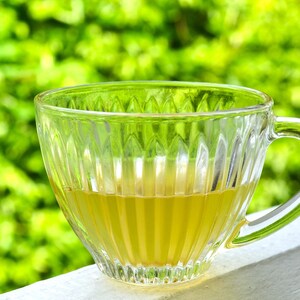 May include: A clear glass teacup with a ribbed design, filled with yellow liquid. The cup has a handle and sits on a white surface, with a blurred green background of foliage. The cup is approximately 3 inches in diameter.