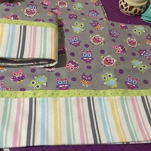 May include: A gray and purple fabric with a pattern of colorful owls and white polka dots. The fabric has a green and white striped border and a pink, yellow, and blue striped border.