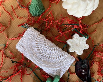 May include: A white crocheted clutch purse, a green Christmas tree, a white snowflake, a white round candle, a green reindeer, and black sunglasses on a red and green shredded paper background.