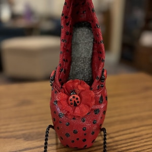 Puede incluir: Una zapatilla de ballet roja y negra con lunares y un adorno de mariquita en una flor. La zapatilla está sobre un soporte de metal negro.