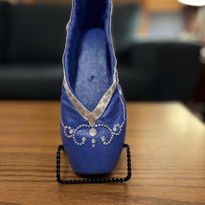 May include: A blue ballet slipper with silver glitter and rhinestones. The slipper is on a black stand.