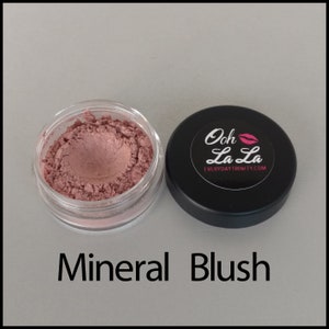 May include: A small, clear jar filled with pink mineral blush next to a black lid. The lid has a white label with the words "Ooh La La" and a pink lip graphic. The text "Mineral Blush" is below the jar.