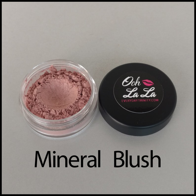 May include: A small, clear jar filled with pink mineral blush next to a black lid. The lid has a white label with the words "Ooh La La" and a pink lip graphic. The text "Mineral Blush" is below the jar.