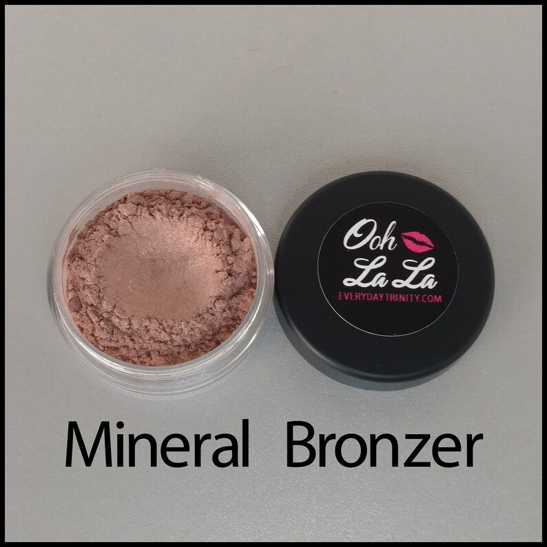 May include: A clear jar filled with mineral bronzer next to a black lid with a white and pink label that reads "Ooh La La" and "EverydayTrinity.com". The text "Mineral Bronzer" is below the jar.
