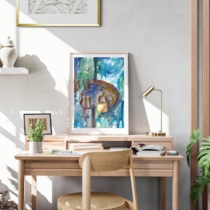 Puede incluir: Una pintura abstracta de acuarela enmarcada con tonos azules, verdes y marrones. La pintura está colgada en una pared blanca sobre un escritorio de madera con una lámpara y una silla.