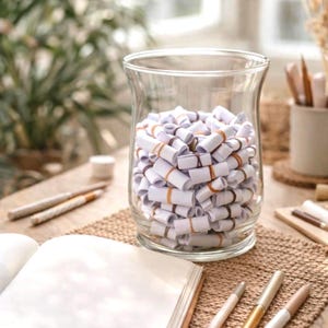 Puede incluir: Un tarro de cristal transparente lleno de rollos de papel blanco asegurados con bandas elásticas naranjas. El tarro está sobre una superficie de madera con un cuaderno, bolígrafos y lápices. La escena está iluminada por luz natural.