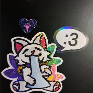May include: A holographic sticker featuring a white cat character holding a light blue water pipe, surrounded by colorful floral shapes. Above the cat is a small heart-shaped sticker, and to the right is a speech bubble sticker with the text :3.