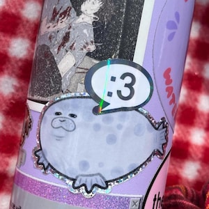 May include: A cylindrical tumbler decorated with various stickers, including a light purple seal with a holographic border and a speech bubble containing the text :3, a black and white illustration of two figures, and text reading the will pass anyway.