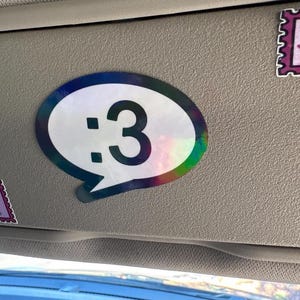May include: A speech bubble sticker with a holographic border and a white center featuring the black text :3. Two additional rectangular stickers are present: one with a lava lamp illustration and another with the text JUST A GIRL.