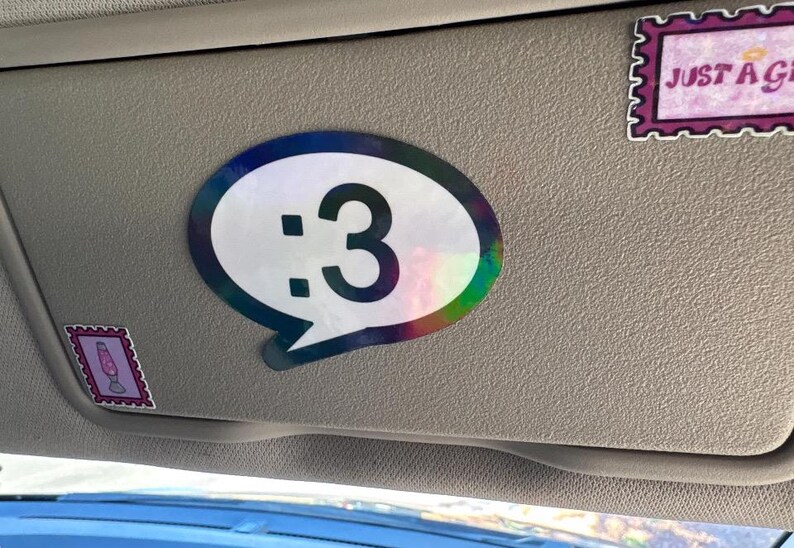 May include: A speech bubble sticker with a holographic border and a white center featuring the black text :3. Two additional rectangular stickers are present: one with a lava lamp illustration and another with the text JUST A GIRL.