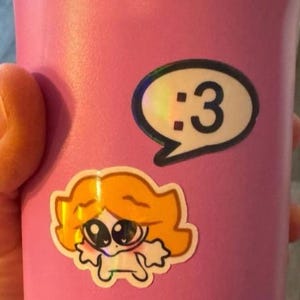 May include: A matte pink tumbler featuring two stickers. One sticker is a white speech bubble containing the black text :3. The second sticker below it depicts a cartoon character with orange hair, large dark eyes, and a small white body.