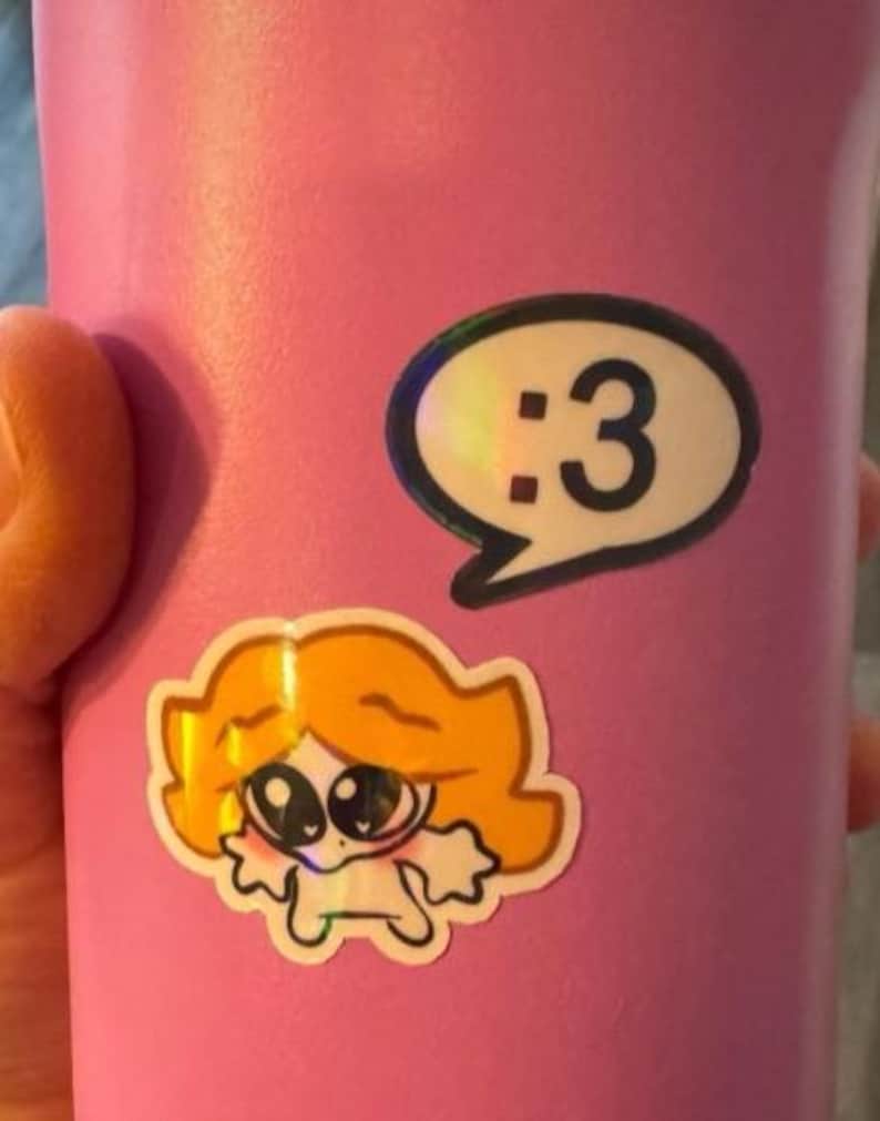 May include: A matte pink tumbler featuring two stickers. One sticker is a white speech bubble containing the black text :3. The second sticker below it depicts a cartoon character with orange hair, large dark eyes, and a small white body.