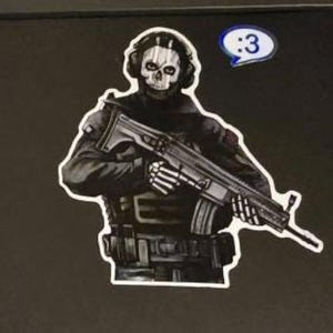 May include: A sticker featuring a character wearing a skull mask and tactical gear, holding a rifle with skeletal hands. The character is dressed in black and grey, and a small blue speech bubble with the text :3 is positioned above the right shoulder.