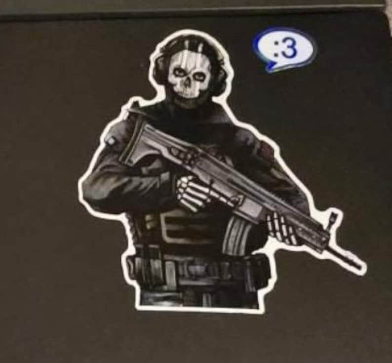May include: A sticker featuring a character wearing a skull mask and tactical gear, holding a rifle with skeletal hands. The character is dressed in black and grey, and a small blue speech bubble with the text :3 is positioned above the right shoulder.
