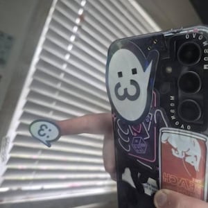 May include: A black smartphone case decorated with multiple stickers, including a large white speech bubble with a :3 emoticon, a neon-style outline of a die, and a rectangular sticker labeled THE PEACH EDITION. A small matching :3 speech bubble sticker is held separately.