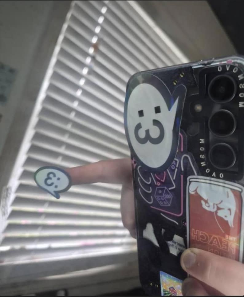 May include: A black smartphone case decorated with multiple stickers, including a large white speech bubble with a :3 emoticon, a neon-style outline of a die, and a rectangular sticker labeled THE PEACH EDITION. A small matching :3 speech bubble sticker is held separately.