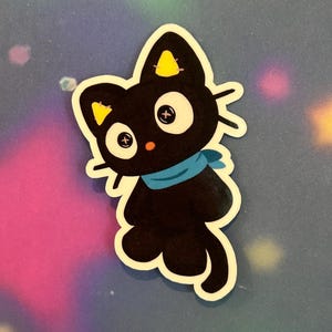 May include: A black cat sticker with white eyes, a blue scarf, and yellow accents on its ears. The sticker has a white border and is set against a colourful, blurred background.