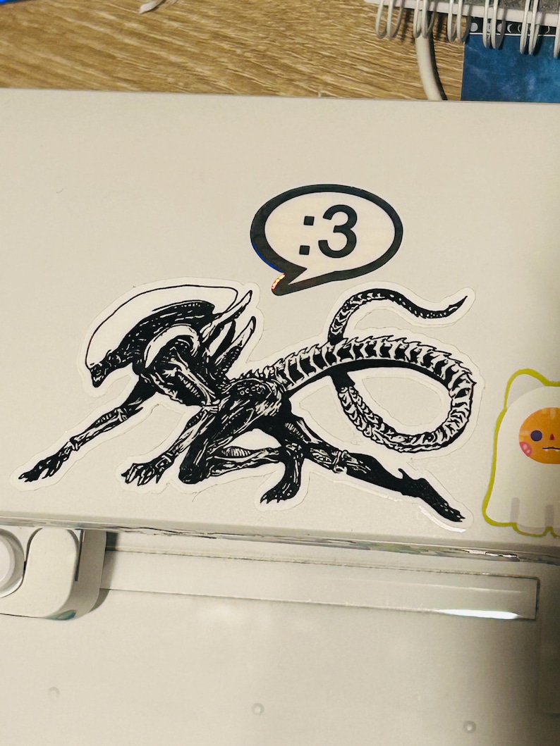 May include: A black and white sticker featuring a detailed illustration of a xenomorph creature in a crouching pose with a long, segmented tail. Above the creature is a speech bubble containing the text :3. A small ghost sticker is visible nearby.
