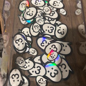 May include: A collection of small, white, speech bubble-shaped stickers with a black outline and the text :3 printed in black. The stickers feature a holographic finish that reflects rainbow colors and are contained within a clear plastic tray.