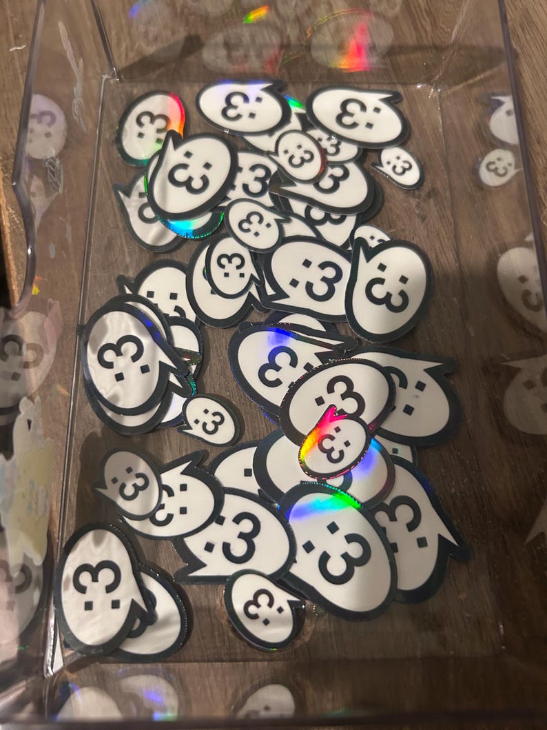 May include: A collection of small, white, speech bubble-shaped stickers with a black outline and the text :3 printed in black. The stickers feature a holographic finish that reflects rainbow colors and are contained within a clear plastic tray.