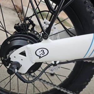 May include: A white bicycle frame section featuring a black and white sticker with a :3 emoticon inside a speech bubble. The frame includes a black rear hub motor, black spokes, a black metal rack, and a black chain. Blue stripes are visible.