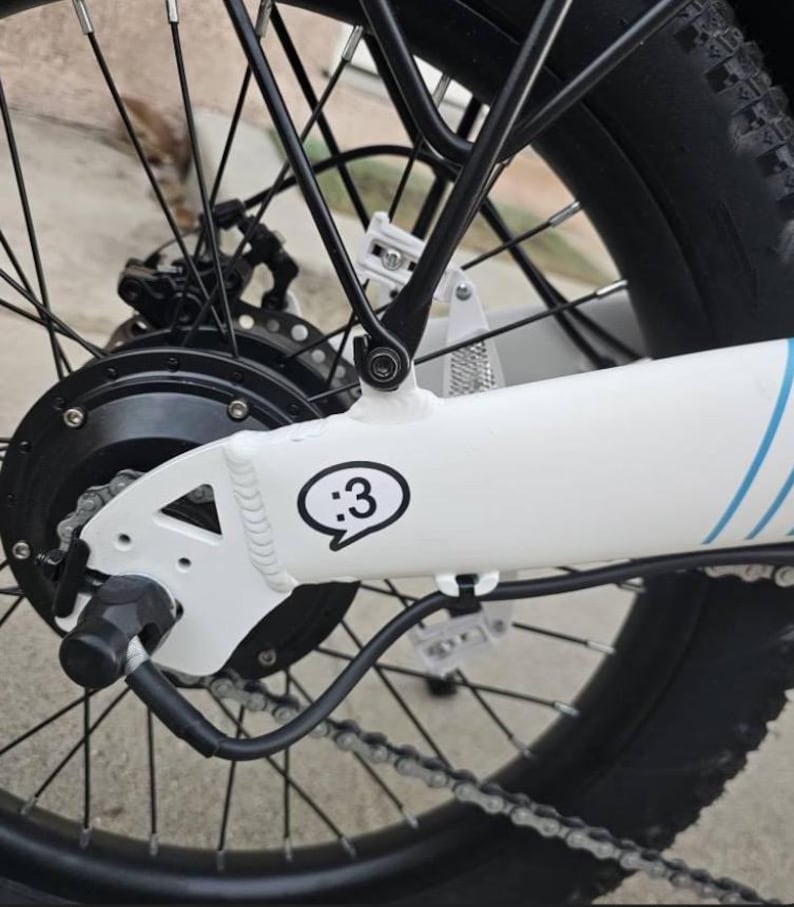 May include: A white bicycle frame section featuring a black and white sticker with a :3 emoticon inside a speech bubble. The frame includes a black rear hub motor, black spokes, a black metal rack, and a black chain. Blue stripes are visible.