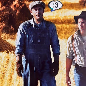 May include: A person wearing a grey flat cap, a blue denim shirt, and blue denim overalls with dark brown work gloves. Beside them, another person wears a black hat and a light shirt. A speech bubble sticker with the text :3 is visible.