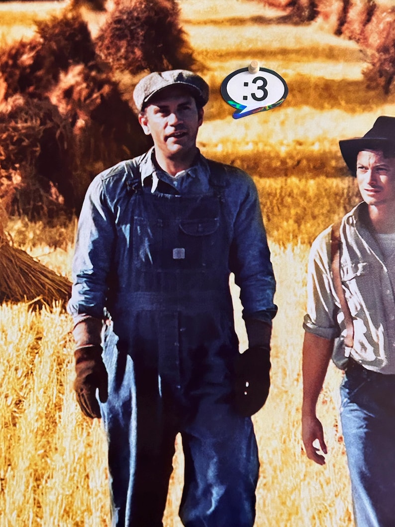 May include: A person wearing a grey flat cap, a blue denim shirt, and blue denim overalls with dark brown work gloves. Beside them, another person wears a black hat and a light shirt. A speech bubble sticker with the text :3 is visible.
