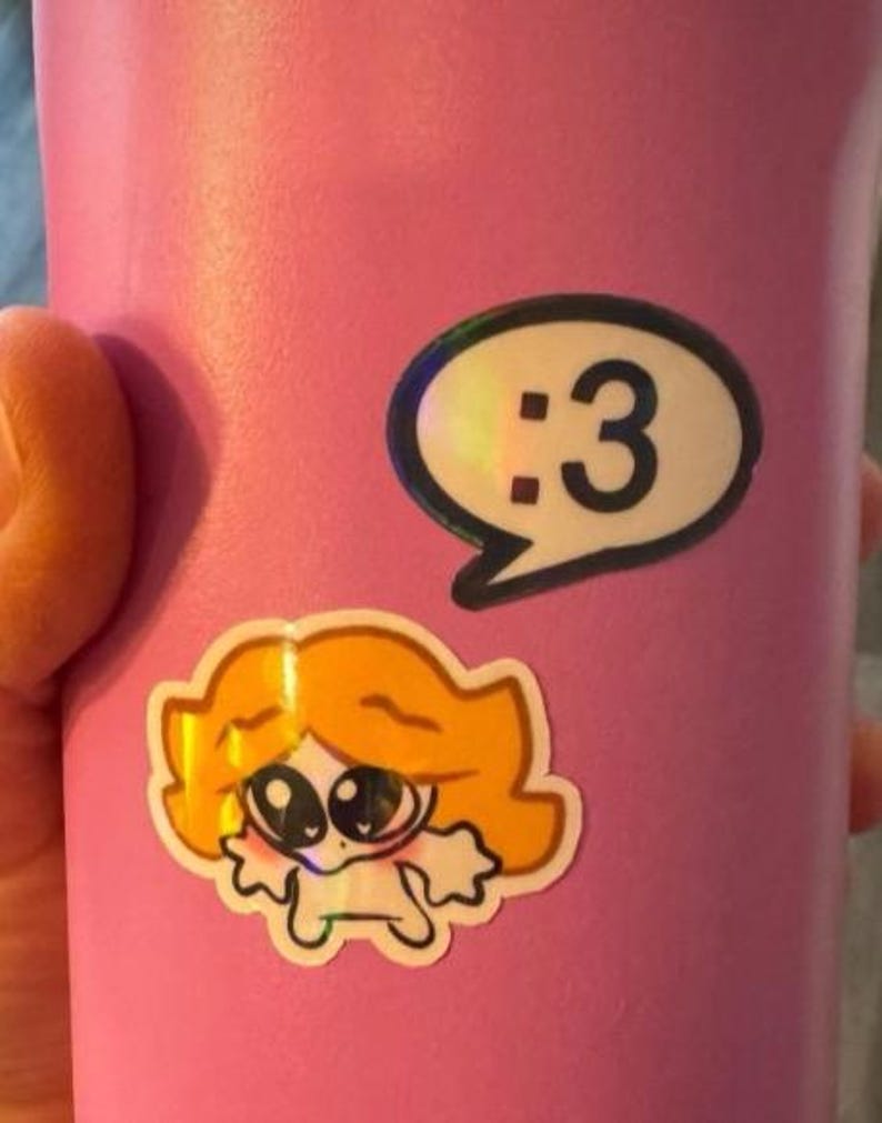 May include: A matte pink tumbler featuring two stickers. One sticker is a white speech bubble containing the black text :3. The second sticker below it depicts a cartoon character with orange hair, large dark eyes, and a small white body.