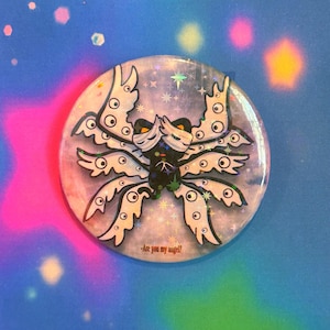 May include: A round button featuring a stylized, white and black creature with multiple eyes and wing-like appendages. The button has a holographic, iridescent finish with the text "-Are you my angel?" at the bottom. The background is a gradient of pink, blue, and purple.