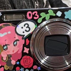 May include: A black digital camera decorated with colorful stickers, including a pink-haired character wearing headphones, a speech bubble with the text :3, the number 03, various stars, and a red rose. The camera features a prominent circular lens.