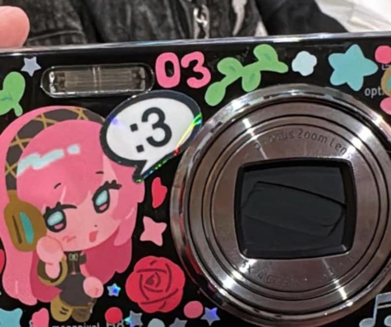 May include: A black digital camera decorated with colorful stickers, including a pink-haired character wearing headphones, a speech bubble with the text :3, the number 03, various stars, and a red rose. The camera features a prominent circular lens.
