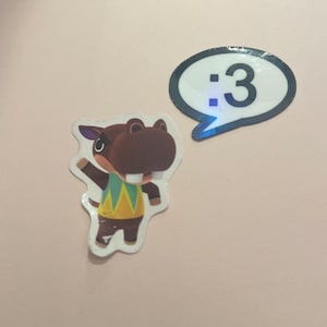 May include: Two stickers featuring a brown hippopotamus character wearing a yellow and green patterned shirt with one arm raised, and a white speech bubble sticker with a black border containing the text :3 in a black font.