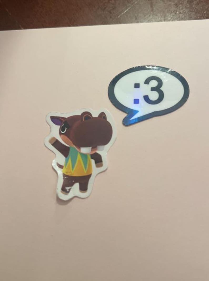 May include: Two stickers featuring a brown hippopotamus character wearing a yellow and green patterned shirt with one arm raised, and a white speech bubble sticker with a black border containing the text :3 in a black font.