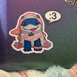 May include: A die-cut sticker featuring a cartoon creature wearing a peach-colored hood with blue accents, a white body covered in red stars, and blue legs. The creature has a wide grin with a visible tongue. A separate speech bubble sticker says :3.