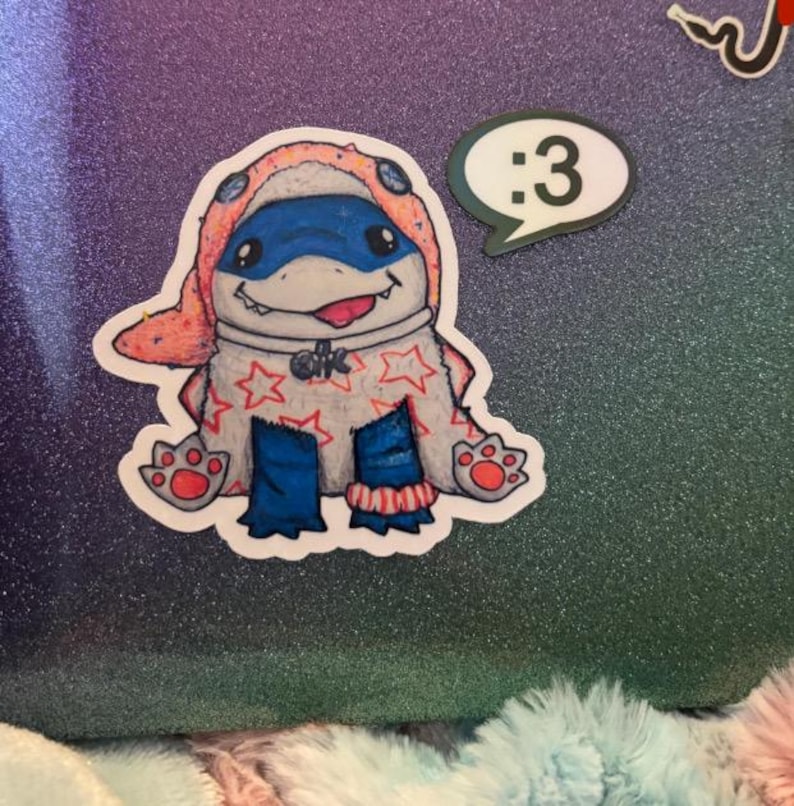 May include: A die-cut sticker featuring a cartoon creature wearing a peach-colored hood with blue accents, a white body covered in red stars, and blue legs. The creature has a wide grin with a visible tongue. A separate speech bubble sticker says :3.