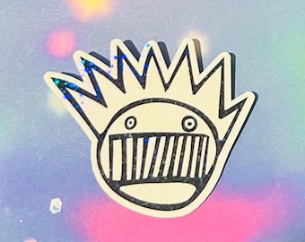 May include: A die-cut sticker featuring a cream-colored circular face with a wide, toothy grin and two small circular eyes. The character has a large, spiky crown-like shape on top. The design includes a dark, glittery finish throughout the surface.