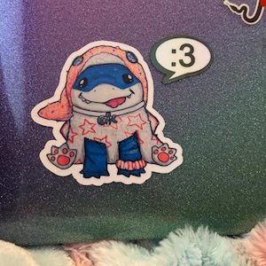 May include: A die-cut sticker featuring a cartoon creature wearing a peach-colored hood with blue accents, a white body covered in red stars, and blue legs. The creature has a wide grin with a visible tongue. A separate speech bubble sticker says :3.