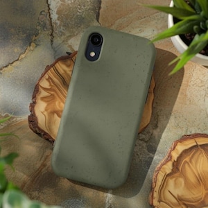 May include: A matte olive green smartphone case with a speckled texture. The case features a single circular camera cutout in the upper left corner with a black lens housing and a small circular flash opening positioned directly below the lens.