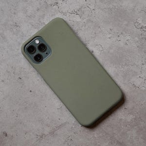 Puede incluir: Funda de tel&eacute;fono verde oliva con acabado mate. La funda est&aacute; dise&ntilde;ada para adaptarse a un tel&eacute;fono inteligente con una c&aacute;mara de triple lente. La funda del tel&eacute;fono est&aacute; sobre una superficie gris texturizada.