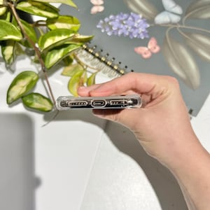 May include: A clear, protective phone case with a transparent design. The case has openings for the charging port and speakers. The phone case is held in a hand, with a blurred background of a notebook and greenery.