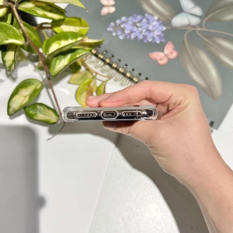 May include: A clear, protective phone case with a transparent design. The case has openings for the charging port and speakers. The phone case is held in a hand, with a blurred background of a notebook and greenery.