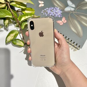 May include: A clear protective phone case on a gold iPhone. The phone has a dual-lens camera and the Apple logo. The case has reinforced corners. The phone is held in a hand.