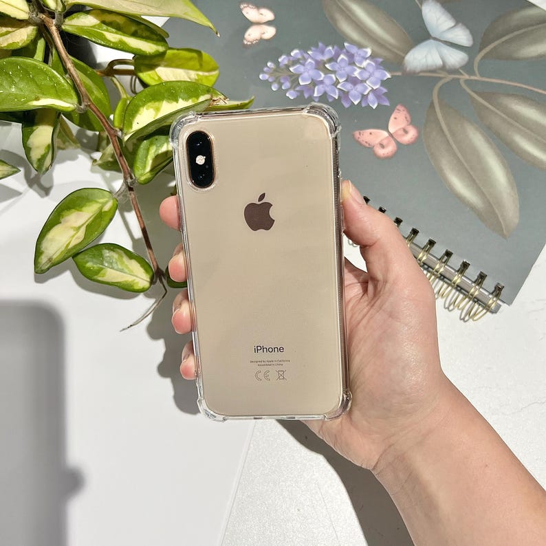 May include: A clear protective phone case on a gold iPhone. The phone has a dual-lens camera and the Apple logo. The case has reinforced corners. The phone is held in a hand.