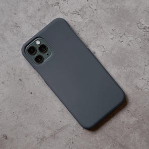 Puede incluir: Una funda de tel&eacute;fono gris oscuro con un recorte para la lente de la c&aacute;mara. La funda est&aacute; hecha de un material liso y cubre la parte trasera y los laterales del tel&eacute;fono. La funda se muestra sobre una superficie gris.