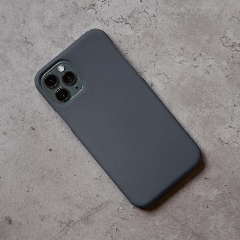 Puede incluir: Una funda de tel&eacute;fono gris oscuro con un recorte para la lente de la c&aacute;mara. La funda est&aacute; hecha de un material liso y cubre la parte trasera y los laterales del tel&eacute;fono. La funda se muestra sobre una superficie gris.