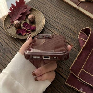 May include: A glossy, dark brown, wavy-edged phone case with a smooth, reflective surface. The case features precise cutouts for the charging port and speakers at the bottom, showcasing a modern, liquid-like design with a deep, rich color tone.
