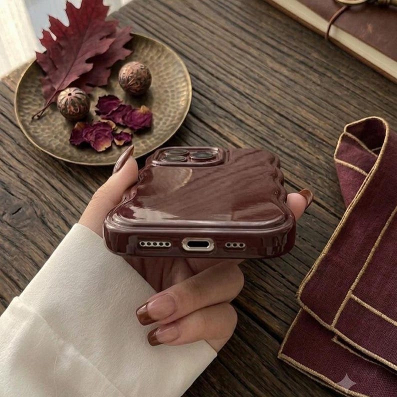 May include: A glossy, dark brown, wavy-edged phone case with a smooth, reflective surface. The case features precise cutouts for the charging port and speakers at the bottom, showcasing a modern, liquid-like design with a deep, rich color tone.