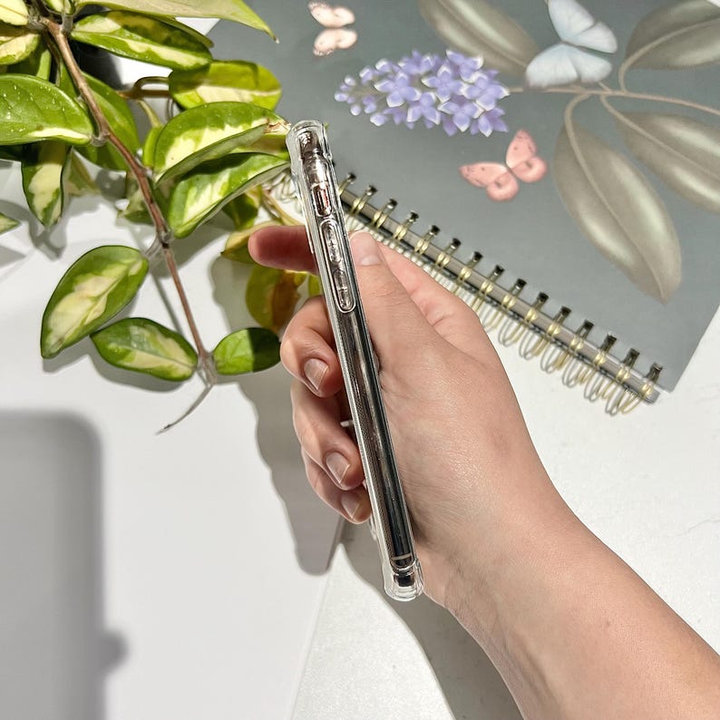 May include: Clear phone case held in hand, showcasing a sleek design. The case is transparent, allowing the phone's color to show through. The image also features a spiral-bound notebook with butterfly and floral illustrations, and a leafy green plant.